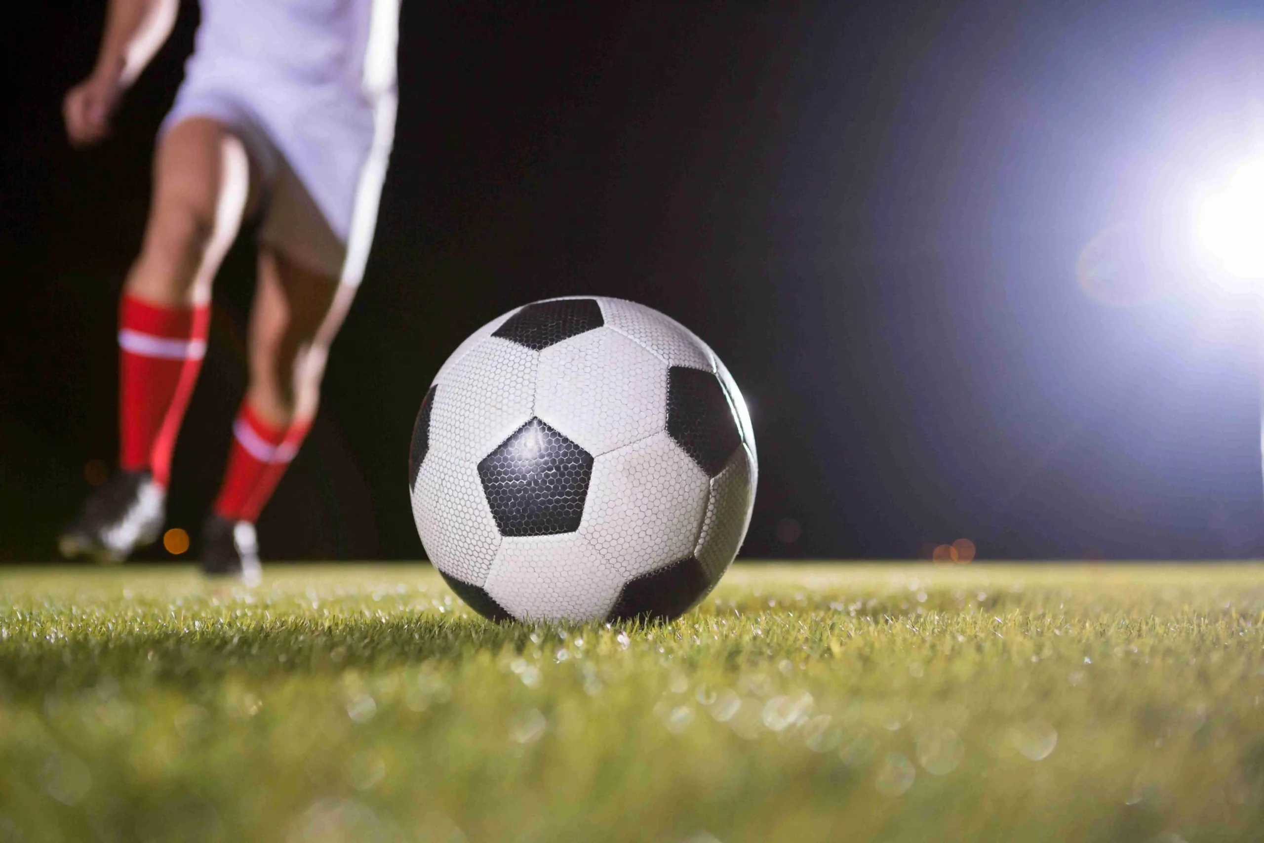 Close-up of a soccer ball on grass at night with a player approaching to kick under stadium floodlights